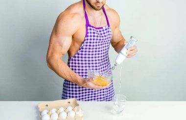 Koruyucu önlüklü şef çırpılmış yumurtaları çırpmak için mutfak masasında çırpıyor. Kaslı adam yemek yapıyor..
