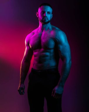 Sportif vücut geliştiren adam renkli neon ışıkta çıplak kas gövdesiyle poz veriyor. Neon stüdyo ışıklarında göğüs ve karın kasları görünüyor..