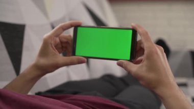 Genç adam kanepede uzanmış cep telefonuna bakıyor, yeşil ekran.