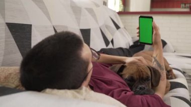 Köpeği kanepede uzanmış, yeşil ekranlı cep telefonuna bakan genç bir adam.