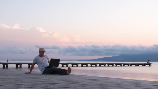 男性旅行者はノートパソコンを使って海岸に座っている デジタル遊牧民またはリモートワーカーの概念 — ストック動画