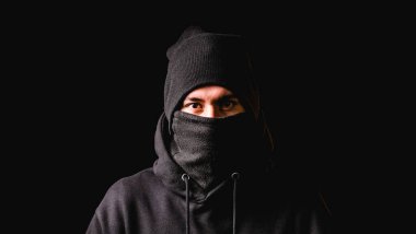 Siyah kar maskeli ve siyah şapkalı bir hırsız karanlık arka planda dururken kameraya bakıyor..