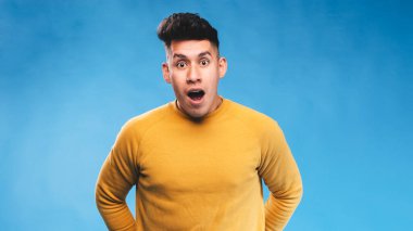 İzole bir arka planda dikilirken yüzünde şok olmuş bir ifadeyle kameraya bakan bir adam..