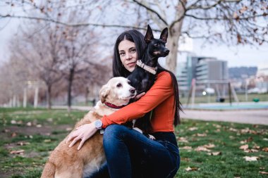 Genç bir kadın parkta köpeklerini tutarken ve kucaklarken kameraya bakıyor..