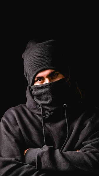 Dieb mit schwarzer Sturmhaube und schwarzer Mütze posiert mit verschränkten Armen vor dunklem Hintergrund. — Stockfoto