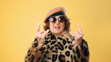 Son sınıf öğrencisi zarif bir kadın sarı arka planda dikilirken parmaklarıyla rock 'n roll hareketi yapıyor..