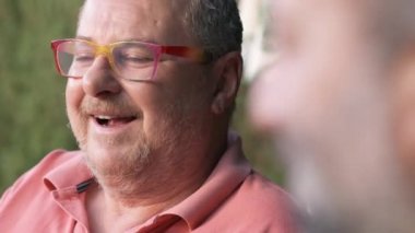 Gözlüklü orta yaşlı erkek yaz günü arka planda bulanık bir şekilde sokakta otururken ekinlere bakan isimsiz bir arkadaşa bakıyor.