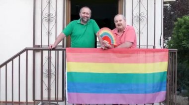 Content homosexual couple looking at camera while standing on balcony of building with symbolic colorful rainbow LGBT flag and fan