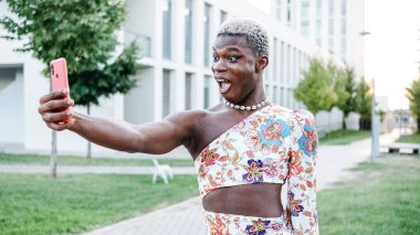 Siyahi ikili olmayan ve Afrikalı genç dışarıda selfie çekiyor. Makyaj yapıyor ve moda kıyafetleri giyiyor.