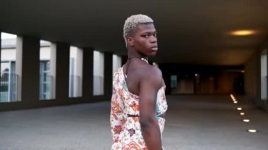 Kendine güvenen ve gülümseyen Afro-Amerikan çift cinsiyetli kısa boyalı saçlı, şık elbiseli ve caddede kameraya bakan kişinin yan görüntüsü.