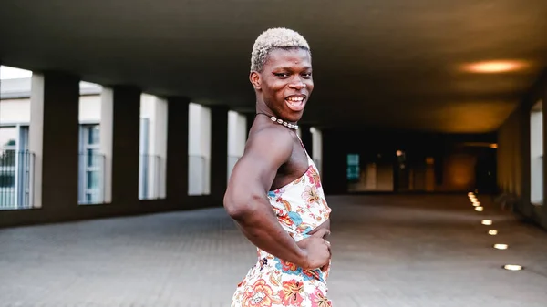 Side view of smiling young African American androgynous person with short dyed hair and makeup in stylish dress standing on street and looking at camera