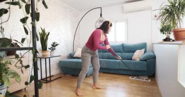 Beyaz kadın müzik dinliyor ve evdeki ahşap zemini temizlerken kulaklıkla şarkı söylüyor. 4k görüntü