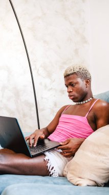 Ciddi bir Afrikalı siyahi kadın kıyafeti giymiş netbook klavyesinde çalışıyor ve daktilo kullanıyor. Evdeki güneşli oturma odasında kanepede oturuyor.