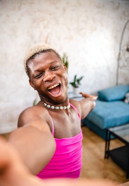 Yukarıdan, pembe elbiseli, saçları boyalı ve dans ederken kameraya bakıp evini gösteren neşeli Afro-Amerikan transseksüel kadından.