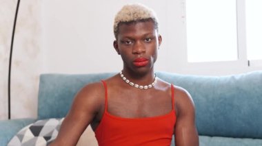 Makyajlı ve kırmızı elbiseli transseksüel bir adam evdeki kanepede otururken kameraya bakıyor..