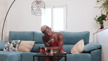 Deniz kabuğu kolyeli Afrikalı bir Amerikalı aynaya bakıyor ve evdeki oturma odasında kanepede otururken göz farı sürüyor.