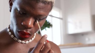 Deniz kabuğundan kolyesi ve kırmızı dudakları olan düşük açılı Afro-Amerikan gay makyajı sırasında evde çizgiler çiziyor.