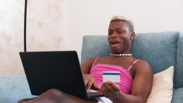 黒アフリカのトランスセクシャル男とともにクレジットカードを入力するラップトップキーボード上でソファに座っている間に注文の支払い自宅でオンラインショッピング — ストック動画