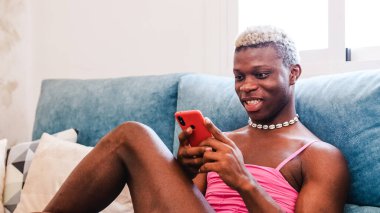 Şık kadın kıyafetleri içinde afro-amerikan erkek akıllı telefondan mesaj atıyor ve evdeki kanepede otururken sesli mesaj dinliyor.