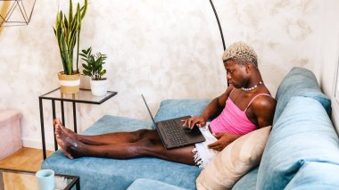 Ciddi Afro-Amerikalı kadın kıyafetleri içinde netbook klavyesinde çalışırken evdeki güneşli oturma odasında kanepede oturuyor.