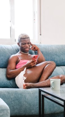 Şık kadın kıyafetleri içinde afro-amerikan erkek akıllı telefondan mesaj atıyor ve evdeki kanepede otururken sesli mesaj dinliyor.