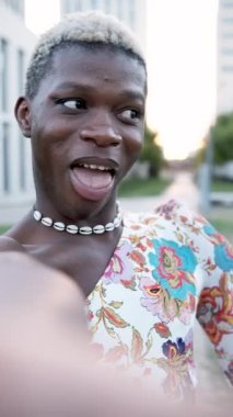 Kısa boyalı saçlı, kadın elbiseli, sokakta selfie çekerken kameraya bakıp poz veren mutlu siyah Afrikalı transseksüel erkek.