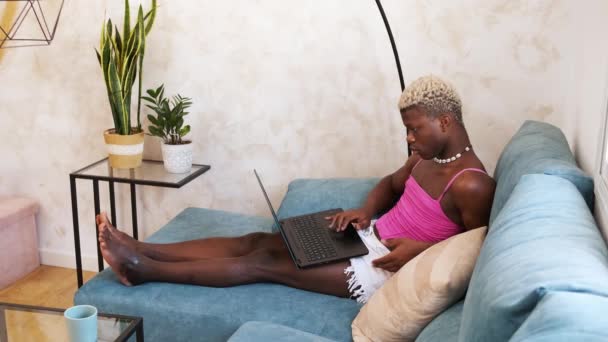 穿着女装的严肃的非洲裔美国人在家里阳光照射的客厅里坐在沙发上 一边工作一边在上网本键盘上打字 — 图库视频影像