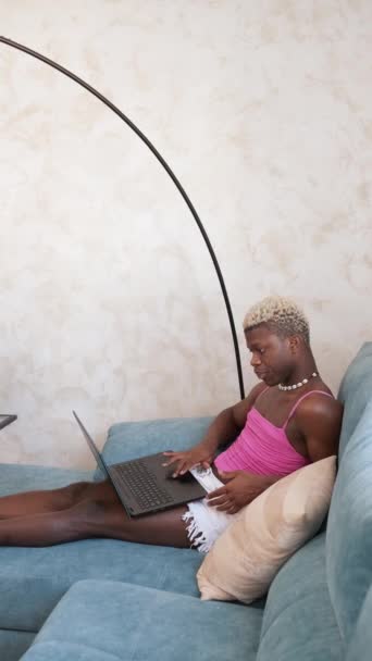 穿着女装的严肃的非洲裔美国人在家里阳光照射的客厅里坐在沙发上 一边工作 一边在上网本键盘上打字 垂直镜头 — 图库视频影像