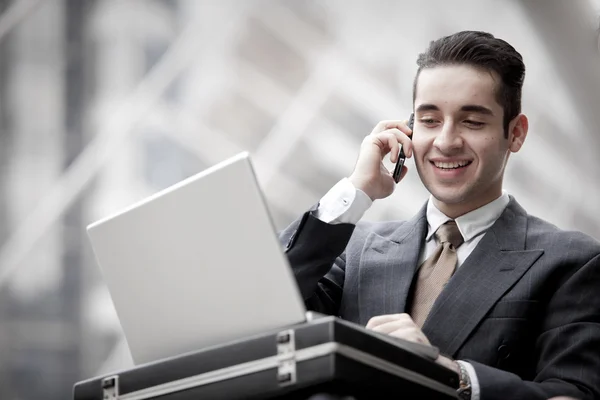 Hombre de negocios guapo que tiene llamada telefónica y el uso de ordenador portátil — Foto de Stock