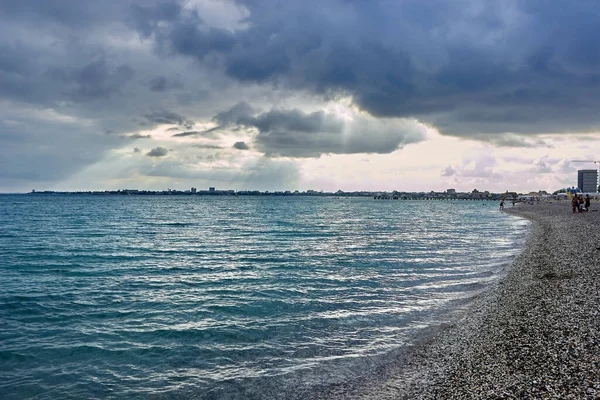 Akşamları küçük dalgalarla mavi deniz. Çakıl taşı plajından su yüzeyinin görüntüsü. İnsanlar kıyıda dinleniyor. Gökyüzündeki kara bulutlar. Doğal deniz manzarası.