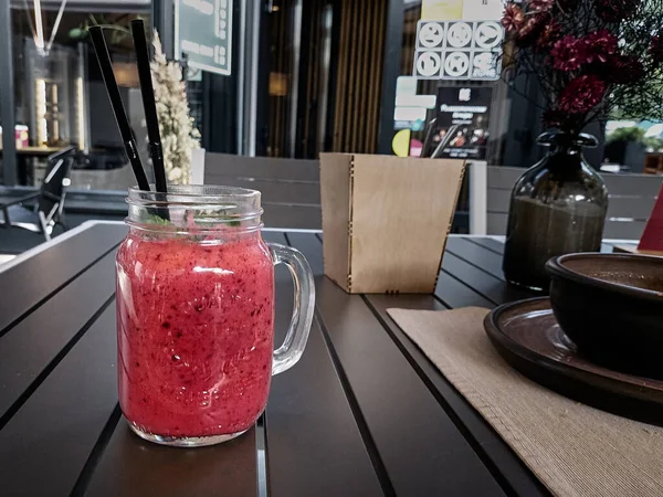 Bright pink berry cocktail or ice cream with mint and a straw in a transparent glass mug on a wood table. Sweet pleasure. Berry smoothie. Cold refreshing dessert in the cafe.