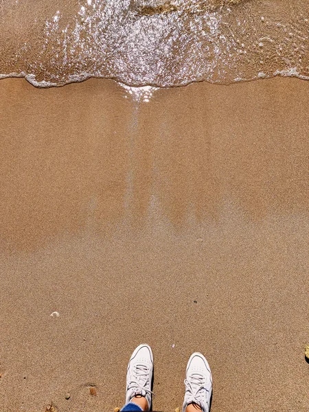 Top view. Woman legs in white sneakers at sea foam waves on sand beach summer day. Ocean clear water seafoam, yellow sand. Summer background. Nature relax concept, holiday vacation. Template for social networks stories. Abstract style for text, desig