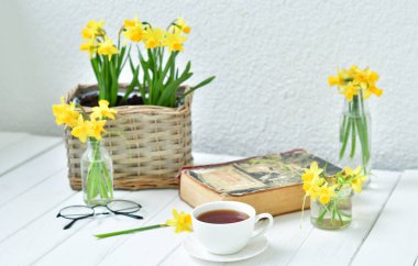 Açılmış eski bir kitap, çay fincanı ve hafif ahşap arka planda sarı nergisler. Samimi bir sabah. Boşluğu kopyala. Seçici odak