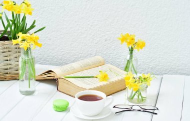 Açılmış eski bir kitap, çay fincanı ve hafif ahşap arka planda sarı nergisler. Samimi bir sabah. Boşluğu kopyala. Seçici odak