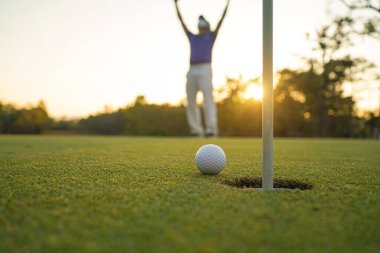 Golfçü topu yeşil golfün üstüne koyuyor, mercek akşam vakti parlıyor. Golfçü vuruşu uzun süre deliğe soktuktan sonra kazanır..                               