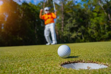 Golfçü topu yeşil golfün üstüne koyuyor, mercek akşam vakti parlıyor. Golfçü vuruşu uzun süre deliğe soktuktan sonra kazanır..                                