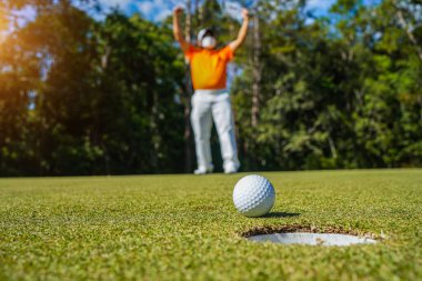 Golfçü topu yeşil golfün üstüne koyuyor, mercek akşam vakti parlıyor. Golfçü vuruşu uzun süre deliğe soktuktan sonra kazanır..                                