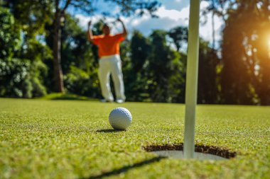 Golfçü topu yeşil golfün üstüne koyuyor, mercek akşam vakti parlıyor. Golfçü vuruşu uzun süre deliğe soktuktan sonra kazanır..                                