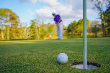 Golfçü topu yeşil golfün üstüne koyuyor, mercek akşam vakti parlıyor. Golfçü vuruşu uzun süre deliğe soktuktan sonra kazanır..                                