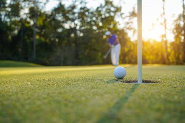 Golfçü topu yeşil golfün üstüne koyuyor, mercek akşam vakti parlıyor. Golfçü vuruşu uzun süre deliğe soktuktan sonra kazanır..                                