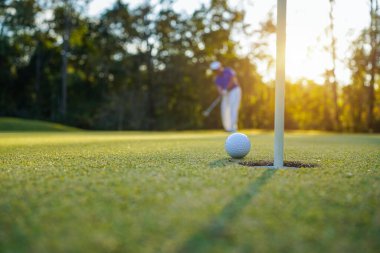 Golfçü topu yeşil golfün üstüne koyuyor, mercek akşam vakti parlıyor. Golfçü vuruşu uzun süre deliğe soktuktan sonra kazanır..                                