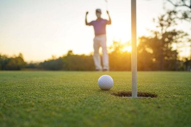 Golfçü topu yeşil golfün üstüne koyuyor, mercek akşam vakti parlıyor. Golfçü vuruşu uzun süre deliğe soktuktan sonra kazanır..                                
