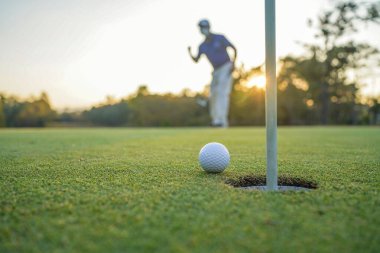Golfçü topu yeşil golfün üstüne koyuyor, mercek akşam vakti parlıyor. Golfçü vuruşu uzun süre deliğe soktuktan sonra kazanır..                                
