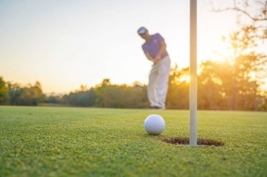Golfçü topu yeşil golfün üstüne koyuyor, mercek akşam vakti parlıyor. Golfçü vuruşu uzun süre deliğe soktuktan sonra kazanır..                                