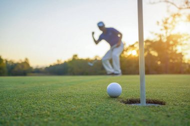 Golfçü topu yeşil golfün üstüne koyuyor, mercek akşam vakti parlıyor. Golfçü vuruşu uzun süre deliğe soktuktan sonra kazanır..                                