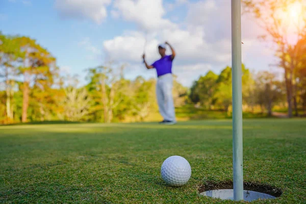 Golfçü topu yeşil golfün üstüne koyuyor, mercek akşam vakti parlıyor. Golfçü vuruşu uzun süre deliğe soktuktan sonra kazanır..                                