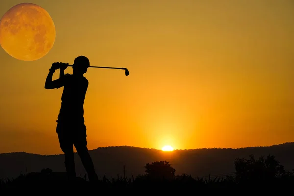 Golfers' hit golf ball toward the hole at sunset silhouetted. Golden morning sky in winter, misty high mountain background.