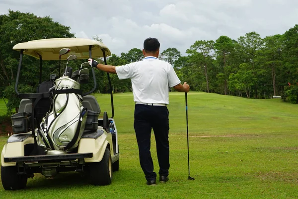 A golfer standing next to a golf cart looks at the golf course to plan the game.