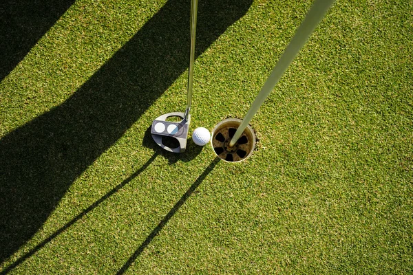 ゴルファーは朝の太陽と美しいゴルフコース上の緑の芝生の上にパターで穴にゴルフボールを置きます — ストック写真