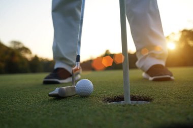 Golfçü topu yeşil golfün üstüne koyuyor, mercek akşam vakti parlıyor. Golfçü vuruşu uzun süre deliğe soktuktan sonra kazanır..                   